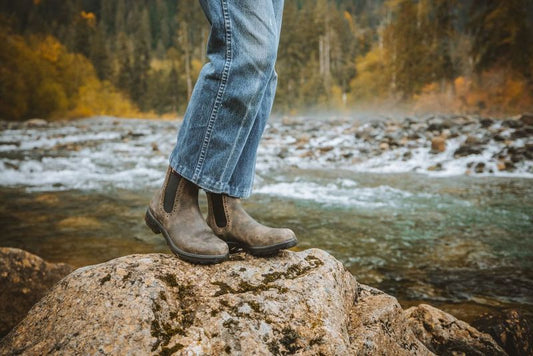 A person wearing a pair of hi top rustic brown blundstones on a rock alongside a river.