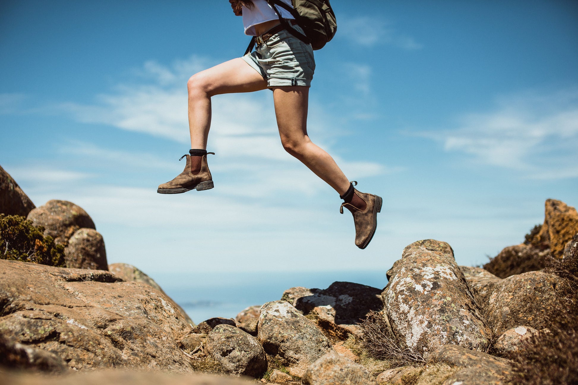 A person wearing rustic brown chisel toe dress blundstones jumping on rocs with a blue sky background.