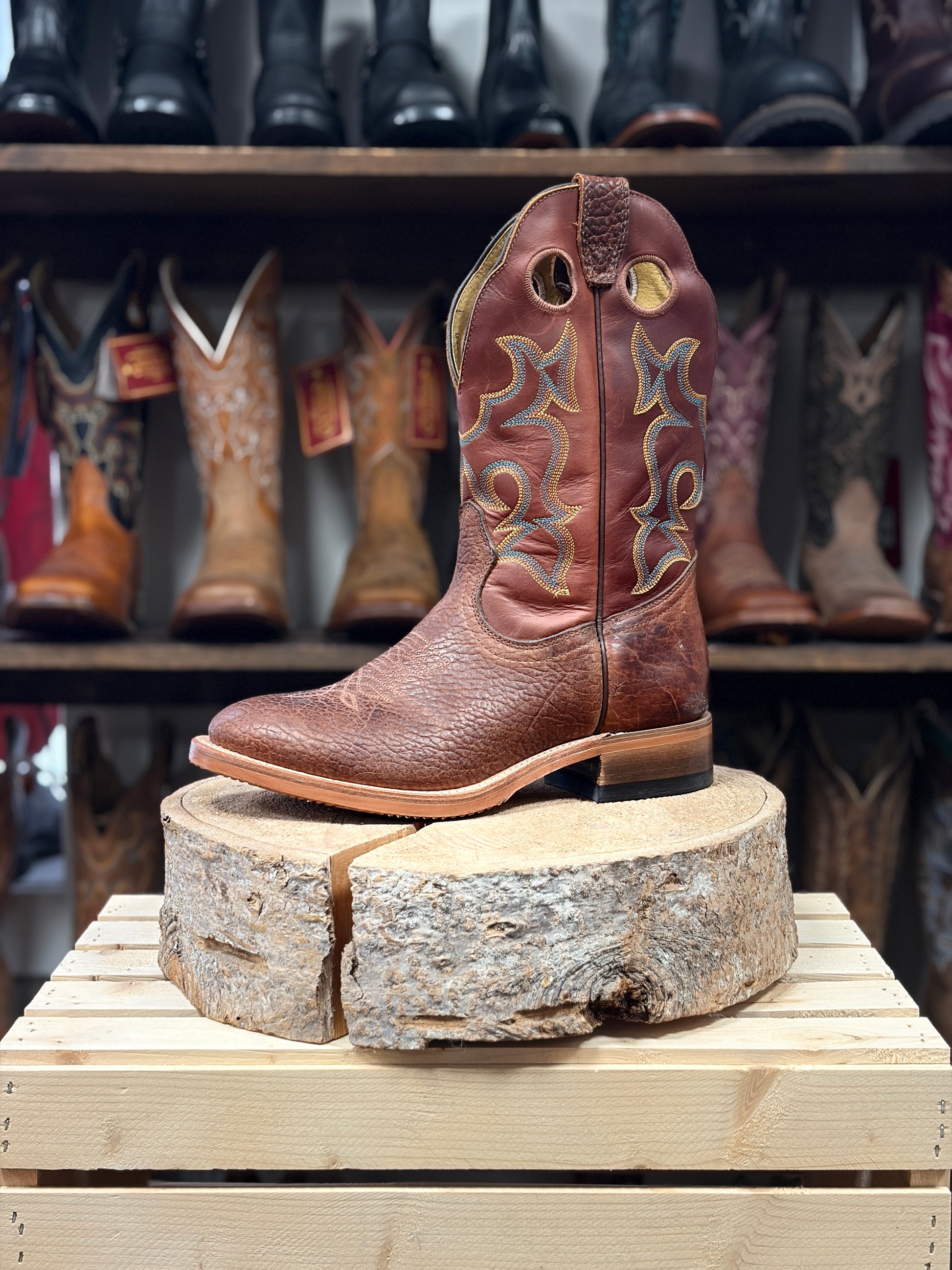 Brown cowboy boot on a wooden block with a background of other boots.