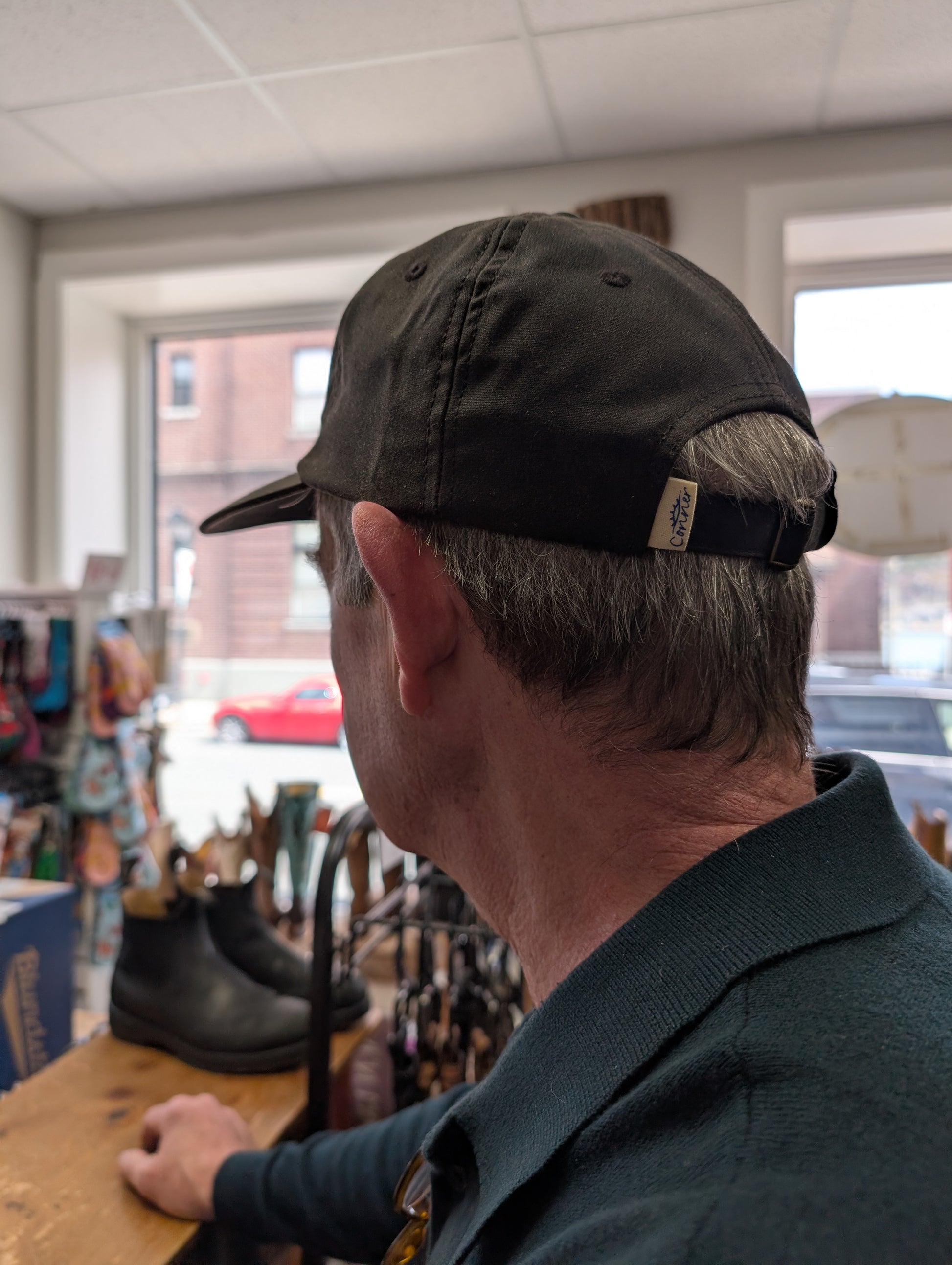 Person wearing a cap in a store setting, highlighting the adjustable band on the back of the dark brown oilskin cap. 